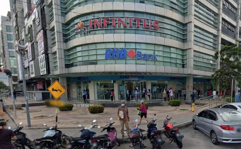Pemandangan aras jalan cawangan RHB Bank yang terletak di sudut bangunan kaca bertingkat moden, dengan papan tanda "Infinitus" kelihatan di tingkat atas. Pejalan kaki berjalan di kaki lima dan beberapa motosikal diletakkan di latar depan