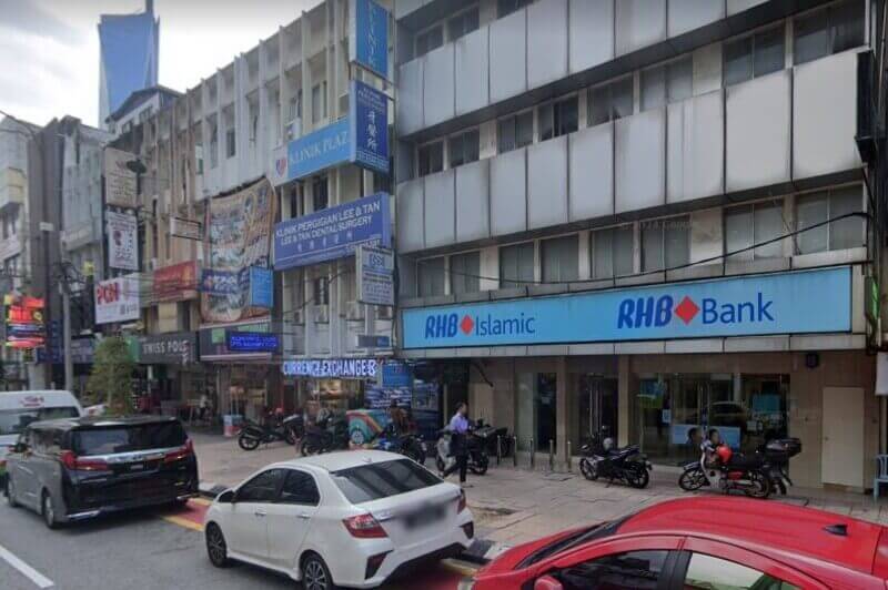 Pemandangan aras jalan luar Bank RHB dan cawangan RHB Islamic di jalan bandar yang sibuk, menampilkan papan tanda biru jernih, kenderaan yang diletakkan dan rumah kedai di sekeliling