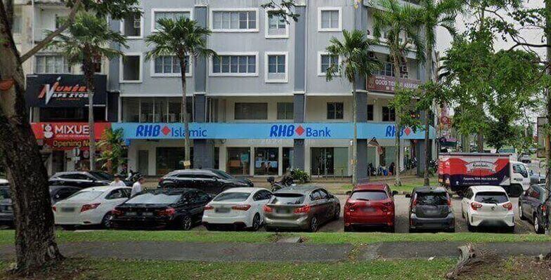Pandangan luar sebuah bank RHB Bank dan cawangan RHB Islamic bertingkat di Kota Kemuning, Shah Alam, yang memaparkan papan tanda kedai biru yang menonjol dan beberapa buah kereta yang diletakkan di tepi jalan di latar depan
