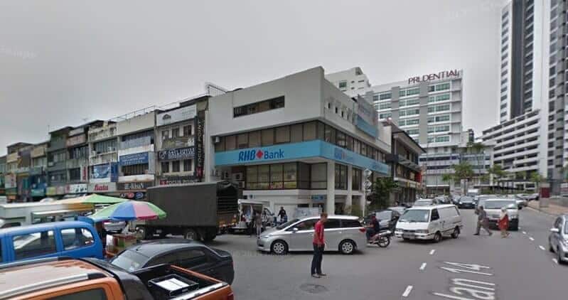 Pemandangan jalan cawangan RHB Bank di selekoh yang sibuk di Seksyen 14, Petaling Jaya, menampilkan bangunan bertingkat putih dengan papan tanda biru, dikelilingi oleh kereta yang diletakkan, penjaja jalanan dan bangunan Prudential di latar belakang