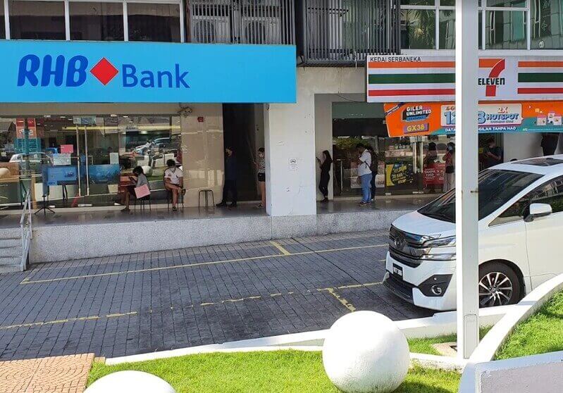 Pemandangan hadapan cawangan RHB Bank dan kedai serbaneka 7-Eleven di deretan rumah kedai dengan beberapa orang beratur di kaki lima