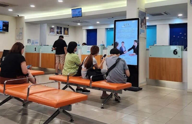 Pemandangan kawasan menunggu di dalam sebuah bank, menunjukkan pelanggan duduk di kerusi oren sementara seorang lagi berdiri di kaunter