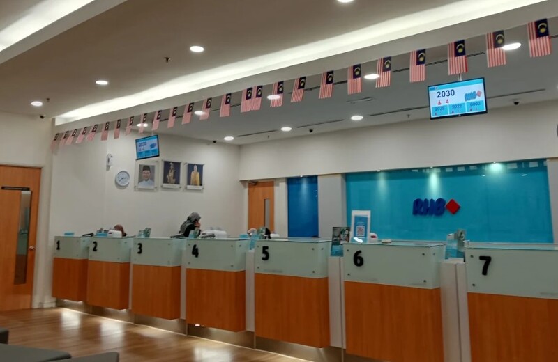 Suasana di dalam cawangan bank RHB, menunjukkan barisan kaunter perkhidmatan bernombor 1 hingga 7 dengan seorang staf di kaunter 4. Siling dihiasi dengan bendera kecil Malaysia